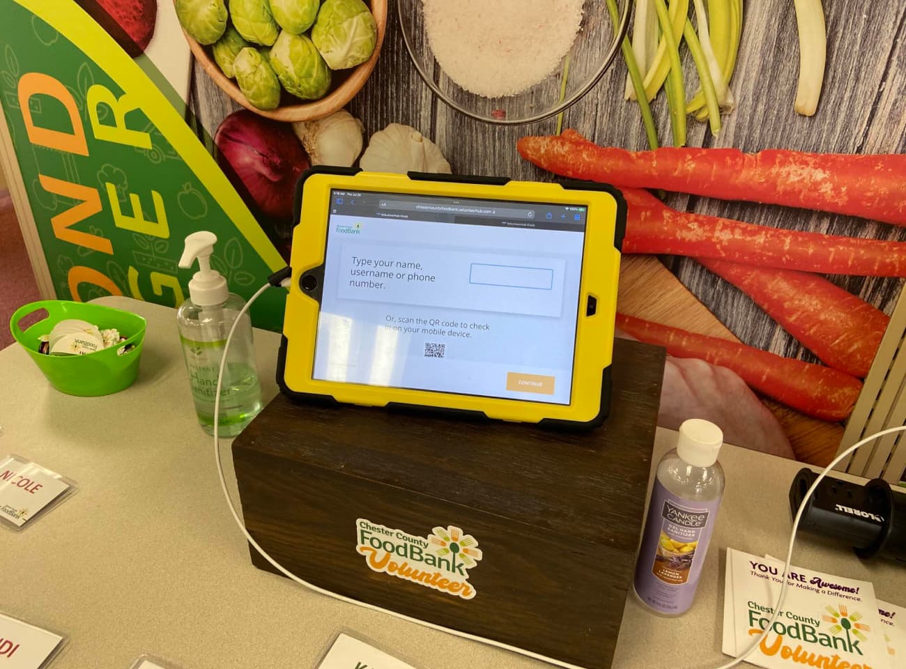 A tablet open to the VolunteerHub mobile kiosk against a backdrop of a mural of vegetables.